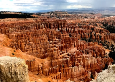 A breathtaking landscape of reddish-brown rock formations known as hoodoos, rising dramatically from the canyon floor. The scene is set under a partly cloudy sky, with layers of rock stretching into the distance and patches of greenery scattered among the formations.
