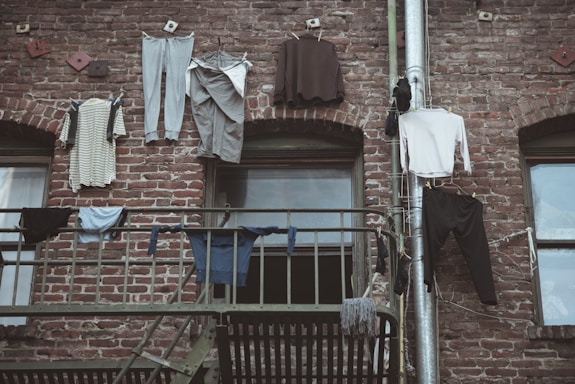 A brick facade with clothing hanging on a clothesline. Several items, including shirts, pants, and socks, are draped over the line and a metal railing, set against the rustic texture of the building's exterior.