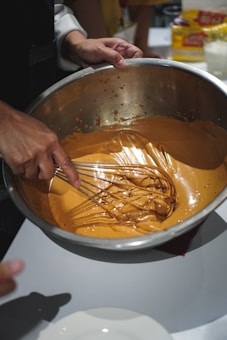 A person whisking a caramel-colored batter in a large metal mixing bowl. The hand is holding a metal whisk, blending the mixture smoothly. There is a white plate and various kitchen items visible in the background.