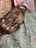A close-up of hands adorned with intricate henna designs, showcasing a ring with a gemstone on one finger. The attire features colorful embroidery with floral patterns in shades of pink, blue, and gold. Multiple pearl bracelets and colorful bangles complement the ensemble.