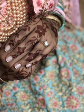 A close-up of hands adorned with intricate henna designs, showcasing a ring with a gemstone on one finger. The attire features colorful embroidery with floral patterns in shades of pink, blue, and gold. Multiple pearl bracelets and colorful bangles complement the ensemble.