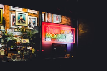 A storefront window displays a variety of framed photographs on wooden shelves, illuminated by a neon sign that reads '1 Hour Photo Processing Fax Copies'. The decor includes plants and small decorative objects, giving a nostalgic vibe. The bright colors of the neon sign contrast with the darker surroundings.