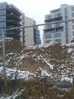 A construction site with a chain-link fence in the foreground and two mid-rise modern apartment buildings in the background. The ground is covered with dirt and patches of snow, suggesting winter or early spring. The buildings feature numerous large windows, and the sky is overcast, adding a gray tone to the scene.