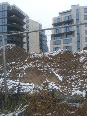 A construction site with a chain-link fence in the foreground and two mid-rise modern apartment buildings in the background. The ground is covered with dirt and patches of snow, suggesting winter or early spring. The buildings feature numerous large windows, and the sky is overcast, adding a gray tone to the scene.