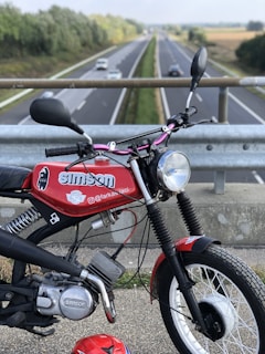A red vintage motorcycle with a 'Simson' logo is parked on an overpass overlooking a highway. The road stretches into the distance, bordered by green trees and fields. Several cars are moving along the highway. The motorcycle features logos and branding, and there is a matching red helmet resting on the ground.