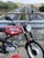 A red vintage motorcycle with a 'Simson' logo is parked on an overpass overlooking a highway. The road stretches into the distance, bordered by green trees and fields. Several cars are moving along the highway. The motorcycle features logos and branding, and there is a matching red helmet resting on the ground.