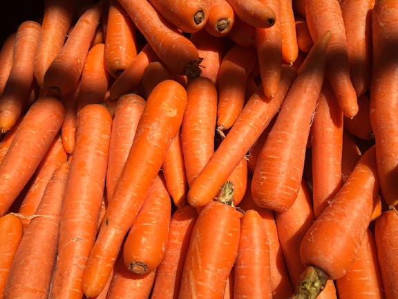 A pile of fresh, unpeeled carrots is closely packed together with visible textures and natural imperfections. The carrots vary slightly in size and shape, and they have a vibrant orange color with a few darker spots on some tips.