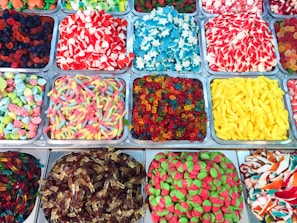An assortment of multi-colored candies displayed in metal trays. The candies come in various shapes such as worms, hearts, stars, and bananas, and are vibrantly colored in reds, yellows, blues, greens, and more. The display is visually appealing with a wide variety of textures.