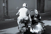 A person rides a scooter with a sidecar along a quiet street. Both rider and passenger are wearing helmets, and the passenger is seated in the sidecar. The street appears empty, with buildings and a tree visible in the background.