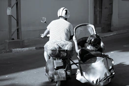 A person rides a scooter with a sidecar along a quiet street. Both rider and passenger are wearing helmets, and the passenger is seated in the sidecar. The street appears empty, with buildings and a tree visible in the background.
