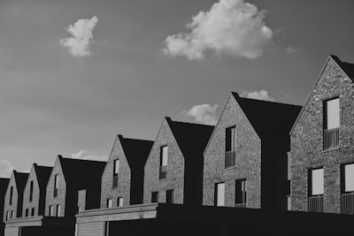 A row of modern brick houses with gabled roofs, set under a sky with scattered clouds. The houses have uniform architecture and appear to be part of a new development or neighborhood.