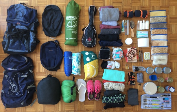 A variety of camping and travel gear is neatly organized on a wooden floor. There are backpacks, a sleeping bag, a tent, rolled-up clothing, toiletries, food items in plastic bags, a hairbrush, a toothbrush, flip-flops, a pair of pink sneakers, containers, and cutlery.