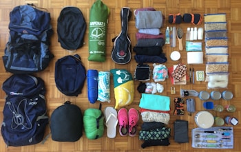 A variety of camping and travel gear is neatly organized on a wooden floor. There are backpacks, a sleeping bag, a tent, rolled-up clothing, toiletries, food items in plastic bags, a hairbrush, a toothbrush, flip-flops, a pair of pink sneakers, containers, and cutlery.