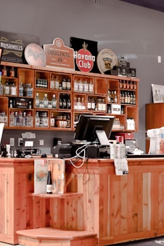 A wooden bar counter is filled with various alcohol bottles arranged neatly on shelves. Behind the counter, there are colorful signs and logos of different liquor brands like Havana Club and Bushmills. The counter also features a cash register, some electronic devices, and promotional materials. The background wall is painted in a muted color, complementing the warm wood tones.