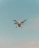 A drone is flying in the center of a clear sky. The drone is white with a camera attached to its underside, indicating it might be used for aerial photography.