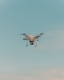 A drone is flying in the center of a clear sky. The drone is white with a camera attached to its underside, indicating it might be used for aerial photography.