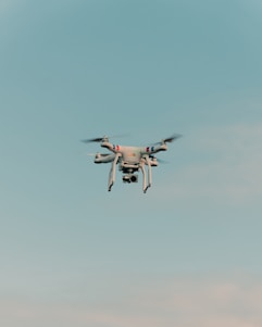 A drone is flying in the center of a clear sky. The drone is white with a camera attached to its underside, indicating it might be used for aerial photography.