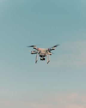 A drone is flying in the center of a clear sky. The drone is white with a camera attached to its underside, indicating it might be used for aerial photography.