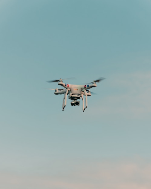 A drone is flying in the center of a clear sky. The drone is white with a camera attached to its underside, indicating it might be used for aerial photography.