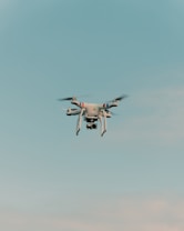 A drone is flying in the center of a clear sky. The drone is white with a camera attached to its underside, indicating it might be used for aerial photography.