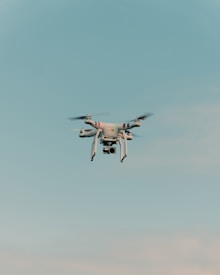 A drone is flying in the center of a clear sky. The drone is white with a camera attached to its underside, indicating it might be used for aerial photography.