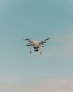 A drone is flying in the center of a clear sky. The drone is white with a camera attached to its underside, indicating it might be used for aerial photography.