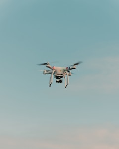 A drone is flying in the center of a clear sky. The drone is white with a camera attached to its underside, indicating it might be used for aerial photography.