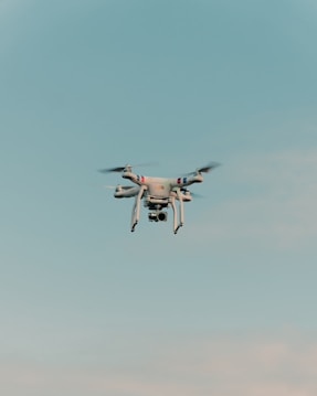 A drone is flying in the center of a clear sky. The drone is white with a camera attached to its underside, indicating it might be used for aerial photography.