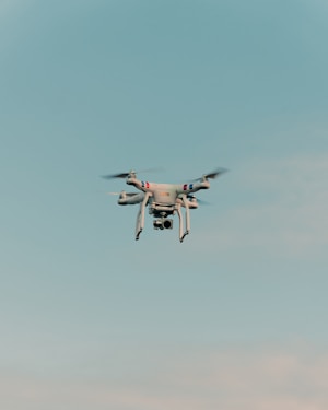 A drone is flying in the center of a clear sky. The drone is white with a camera attached to its underside, indicating it might be used for aerial photography.