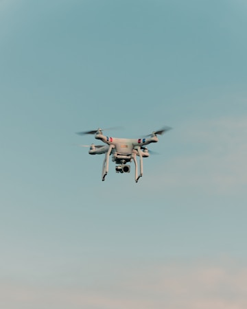 A drone is flying in the center of a clear sky. The drone is white with a camera attached to its underside, indicating it might be used for aerial photography.
