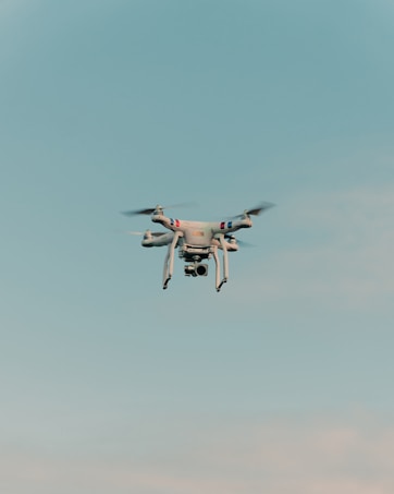 A drone is flying in the center of a clear sky. The drone is white with a camera attached to its underside, indicating it might be used for aerial photography.