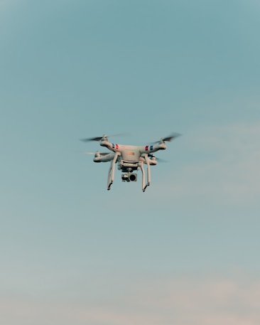 A drone is flying in the center of a clear sky. The drone is white with a camera attached to its underside, indicating it might be used for aerial photography.