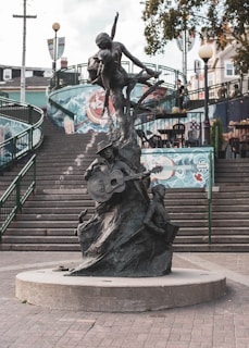 A public sculpture situated in a square, showing a musician playing a guitar and a child playing an accordion. The sculpture is crafted with an artistic, flowing design and is placed on a circular concrete base. Behind it, there is a staircase leading upwards, with colorful murals on the walls alongside the steps. There are green metal railings on the sides and a few streetlights and trees in the background.