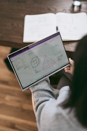 A person is holding a laptop displaying geometric diagrams and equations. The screen shows a cube, a pyramid, and a cylinder with annotations. In the background, there is an open notebook on a wooden table, suggesting a study or learning environment.