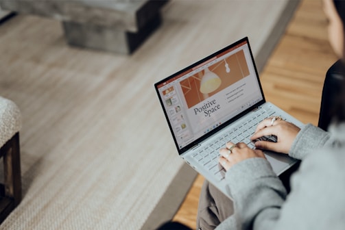 A person is using a laptop with a presentation slide open on the screen. The slide is titled 'Positive Space' and features a graphic of a lamp. The individual is seated in a cozy environment with a carpeted floor and a wooden table nearby.