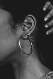 A close-up of a person's ear adorned with several piercings, including a large hoop earring and two smaller studs. The ear is highlighted against a dark background, and a hand is visible in the upper right corner. The image is in black and white, emphasizing texture and contrast.