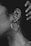 A close-up of a person's ear adorned with several piercings, including a large hoop earring and two smaller studs. The ear is highlighted against a dark background, and a hand is visible in the upper right corner. The image is in black and white, emphasizing texture and contrast.