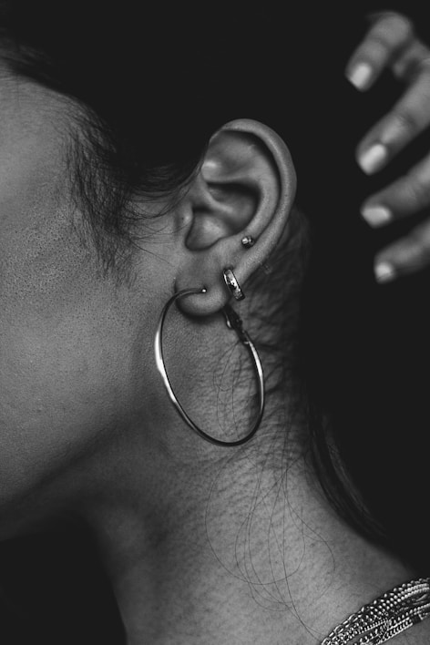 A close-up of a person's ear adorned with several piercings, including a large hoop earring and two smaller studs. The ear is highlighted against a dark background, and a hand is visible in the upper right corner. The image is in black and white, emphasizing texture and contrast.
