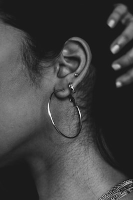 A close-up of a person's ear adorned with several piercings, including a large hoop earring and two smaller studs. The ear is highlighted against a dark background, and a hand is visible in the upper right corner. The image is in black and white, emphasizing texture and contrast.