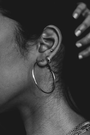 A close-up of a person's ear adorned with several piercings, including a large hoop earring and two smaller studs. The ear is highlighted against a dark background, and a hand is visible in the upper right corner. The image is in black and white, emphasizing texture and contrast.