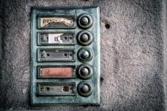 An old, weathered doorbell panel mounted on a textured stone wall. The panel has multiple round buttons arranged vertically, with fading and peeling labels next to each one. The buttons appear tarnished and worn from use.