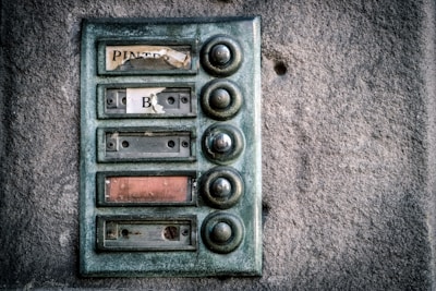 An old, weathered doorbell panel mounted on a textured stone wall. The panel has multiple round buttons arranged vertically, with fading and peeling labels next to each one. The buttons appear tarnished and worn from use.