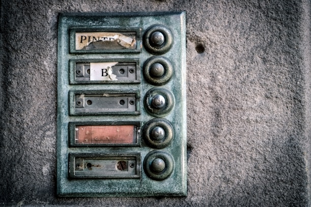 An old, weathered doorbell panel mounted on a textured stone wall. The panel has multiple round buttons arranged vertically, with fading and peeling labels next to each one. The buttons appear tarnished and worn from use.