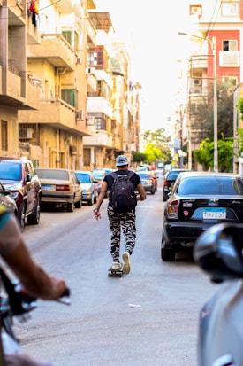 A person wearing camouflage pants and a backpack is skateboarding down a narrow urban street. Surrounding them are parked cars and residential buildings, with balconies and some greenery. The scene suggests a lively, everyday urban environment.