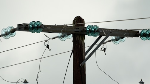 A wooden utility pole supports several electrical insulators made of glass, which are arranged in a circular pattern at the top of the pole. The glass insulators are a light blue-green color, and several wires are attached to them, extending outwards. The background is a cloudy gray sky.
