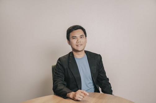 A person is seated at a round wooden table, wearing a blue shirt and a black blazer. The background is plain and neutral, creating a simple and professional atmosphere.