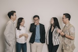 A group of five adults standing together casually, engaging in a lively conversation and sharing smiles. They are dressed in smart casual attire, with varying patterns and colors in their clothing. The setting is simple, with a plain off-white background.