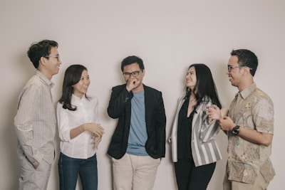 A group of five adults standing together casually, engaging in a lively conversation and sharing smiles. They are dressed in smart casual attire, with varying patterns and colors in their clothing. The setting is simple, with a plain off-white background.