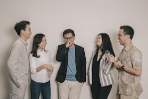 A group of five adults standing together casually, engaging in a lively conversation and sharing smiles. They are dressed in smart casual attire, with varying patterns and colors in their clothing. The setting is simple, with a plain off-white background.