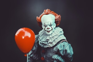 A creepy clown with a white face, red nose, and exaggerated makeup holds a bright red balloon. The clown has orange hair and wears a ruffled gray outfit, set against a dark background.